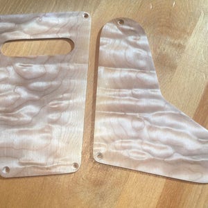 May include: Two wooden guitar pickguards with a light wood grain pattern. The pickguards have multiple holes for mounting screws.