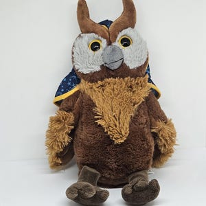 May include: A plush owl toy with brown, tan, and gray coloring. The owl has large yellow eyes, a gray beak, and a fluffy chest. It wears a dark blue cape with a gold trim and white stars. The owl is sitting upright.