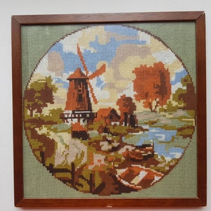 May include: A framed cross-stitch embroidery depicting a windmill in a rural landscape. The scene features a windmill with a red roof and white walls, surrounded by green trees and a blue sky with white clouds. A small river flows through the scene, and there are two boats in the water.
