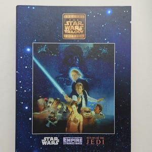 Puede incluir: Una caja rectangular de Star Wars Trilogy Special Edition con un fondo espacial azul oscuro lleno de estrellas. La portada muestra una ilustración central de Luke Skywalker, Han Solo, la Princesa Leia y Darth Vader, junto a los títulos de las películas.