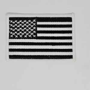 Peut inclure: Patch brodé du drapeau américain en noir et blanc. Le patch a une bordure blanche et est fait de tissu.