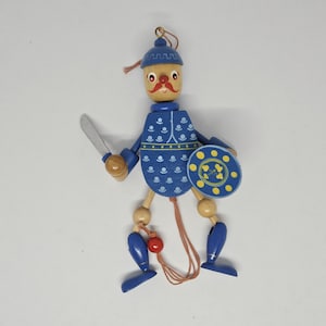 May include: A blue wooden puppet with a yellow shield, a red bead, and a sword. The puppet has a painted face with a mustache and a red hat.