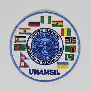 Könnte beinhalten: Ein weißer runder Patch mit einem blauen Rand. In der Mitte des Patches befindet sich ein blauer Kreis mit einem weißen UN-Logo. Der Kreis ist umgeben von einem weißen Ring mit dem Text "United Nations Mission in Sierra Leone" in blau. Der Patch ist umgeben von einem Ring aus Flaggen verschiedener Länder, darunter Russland, Nigeria, Ghana, Indien, Palästina, Nepal, Ukraine, Pakistan, Uganda und Sambia.