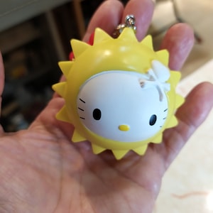 May include: A yellow sun-shaped keychain with a white Hello Kitty face in the center. The keychain has a red bow on top and a white flower on the side.