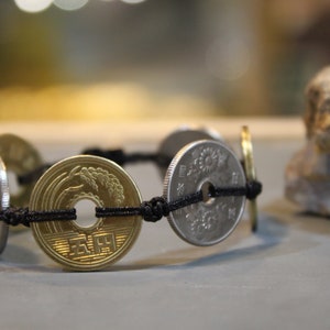 May include: A black cord bracelet with five silver and gold Japanese yen coins. The coins are tied together with a black cord and have a floral design.