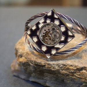 Può includere: Un braccialetto d'argento con un ciondolo quadrato che presenta un design geometrico bianco e viola con un meccanismo di orologeria visibile.