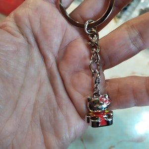 Peut inclure: Porte-clés argenté avec un charme Hello Kitty rouge, jaune et noir. Le charme est une figurine 3D d'Hello Kitty portant une robe rouge et un nœud blanc.