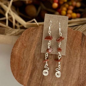 May include: A pair of dangling earrings featuring silver-toned wire wrapped around small, irregular, shimmering reddish-brown goldstone chips. Each earring terminates in a coiled wire spiral at the bottom, suspended from a simple silver-toned hook.