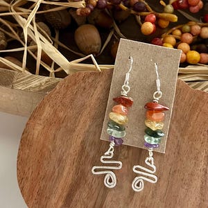 May include: A pair of dangle earrings featuring a vertical stack of multi-colored chip beads in red, orange, yellow, green, and purple, suspended from silver-toned metal hooks. Below the beads, a silver-toned wire is bent into a swirling, abstract shape.
