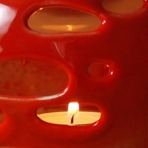 May include: A red ceramic candle holder with a cut-out design. A white candle is burning inside the holder.