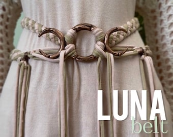 Puede incluir: Un cinturón trenzado con una hebilla de cobre en forma de media luna. El cinturón está hecho de dos tonos de tela, un beige claro y un verde claro. El cinturón se llama "LUNA belt HALF BRAIDED style".