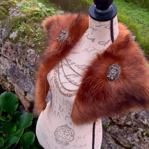 May include: A brown faux fur shawl with two decorative metal brooches. The shawl is displayed on a mannequin with a white background.