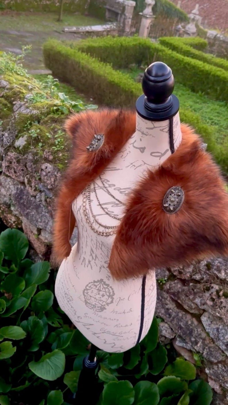 May include: A brown faux fur shawl with two decorative metal brooches. The shawl is displayed on a mannequin with a white background.