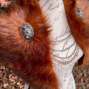 May include: A brown faux fur collar with two silver metal embellishments. The collar is displayed on a mannequin with a white background.