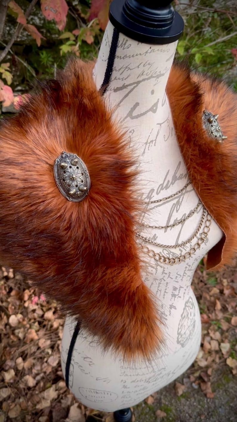 May include: A brown faux fur collar with two silver metal embellishments. The collar is displayed on a mannequin with a white background.