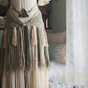 May include: A cream-colored tiered dress featuring a sage green macrame hip belt with long fringe tassels and a central dark triangular knot ornament. The belt is constructed from braided cord and includes a long hanging tie with a wooden bead accent.