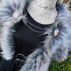 May include: A black faux fur collar with a silver chain necklace and two silver circular brooches. The collar is on a black shirt with a silver belt.