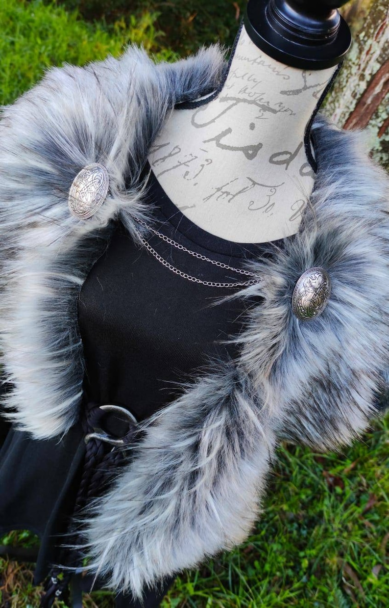 May include: A black faux fur collar with a silver chain necklace and two silver circular brooches. The collar is on a black shirt with a silver belt.