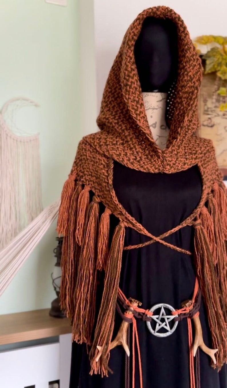 May include: A brown hooded shawl with fringe, worn over a black dress with a pentagram belt buckle and antler accents.