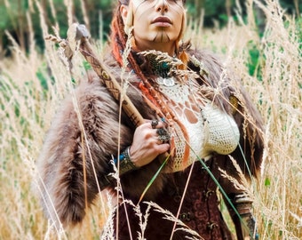 May include: A woman in a forest setting, wearing a brown leather corset, a fur-trimmed jacket, and a beaded headpiece. She is holding a wooden staff with a metal head.