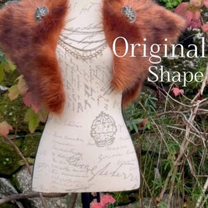 May include: A brown faux fur shawl with two silver brooches. The shawl is draped over a white mannequin with a black stand. The mannequin is standing in front of a stone wall with green plants.