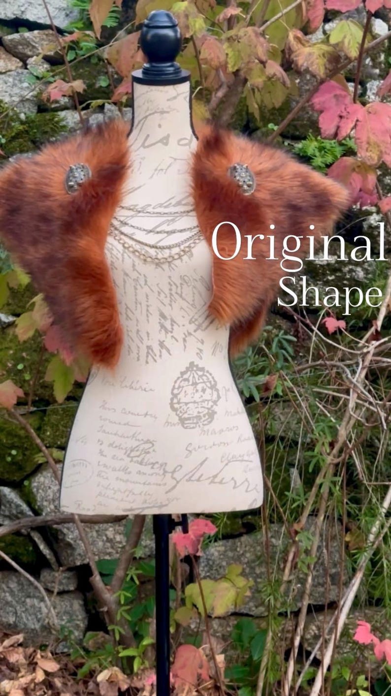 May include: A brown faux fur shawl with two silver brooches. The shawl is draped over a white mannequin with a black stand. The mannequin is standing in front of a stone wall with green plants.