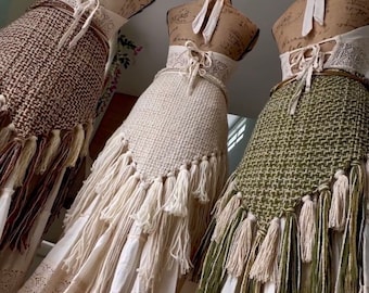 May include: Three crocheted skirts with fringe, each in a different color: brown, white, and green. The skirts are displayed on mannequins and have a white lace trim.