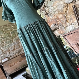 May include: A long, tiered, green cotton dress with a square neckline and short, ruffled sleeves. The dress is photographed on a mannequin against a stone wall.
