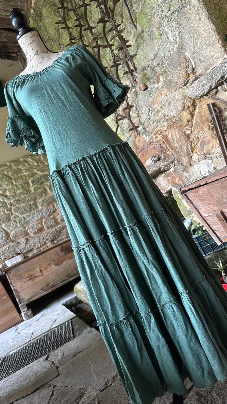 May include: A long, tiered, green cotton dress with a square neckline and short, ruffled sleeves. The dress is photographed on a mannequin against a stone wall.