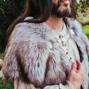 May include: A person wearing a white shirt with a fur collar and silver chain with decorative pendants. The fur collar is white and gray.