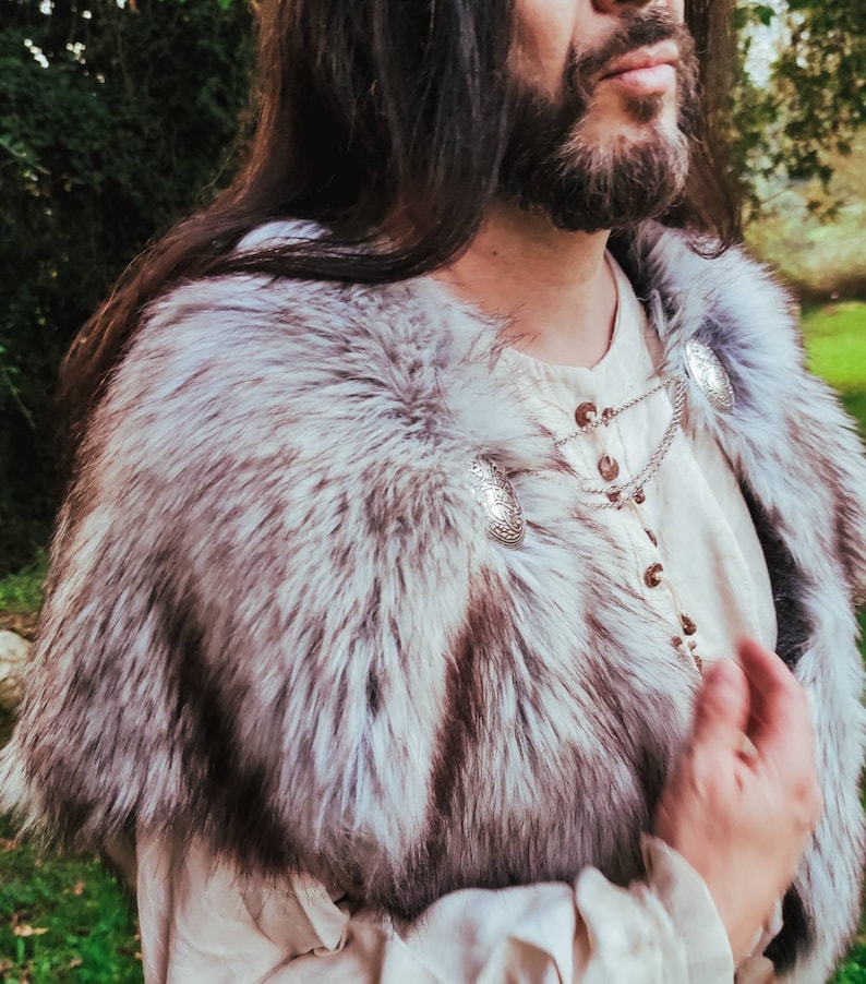 May include: A person wearing a white shirt with a fur collar and silver chain with decorative pendants. The fur collar is white and gray.