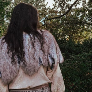May include: A person wearing a white shirt and a fur shawl. The shawl is made of white and gray fur and has a long, flowing design. The person is standing in a forest setting.