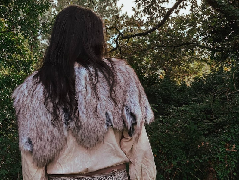 May include: A person wearing a white shirt and a fur shawl. The shawl is made of white and gray fur and has a long, flowing design. The person is standing in a forest setting.