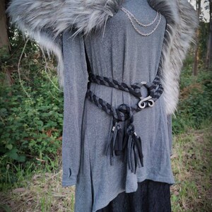 May include: A gray long-sleeved tunic with a black braided belt and a silver buckle. The tunic has a faux fur collar with a silver brooch. The model is wearing a black skirt underneath the tunic.