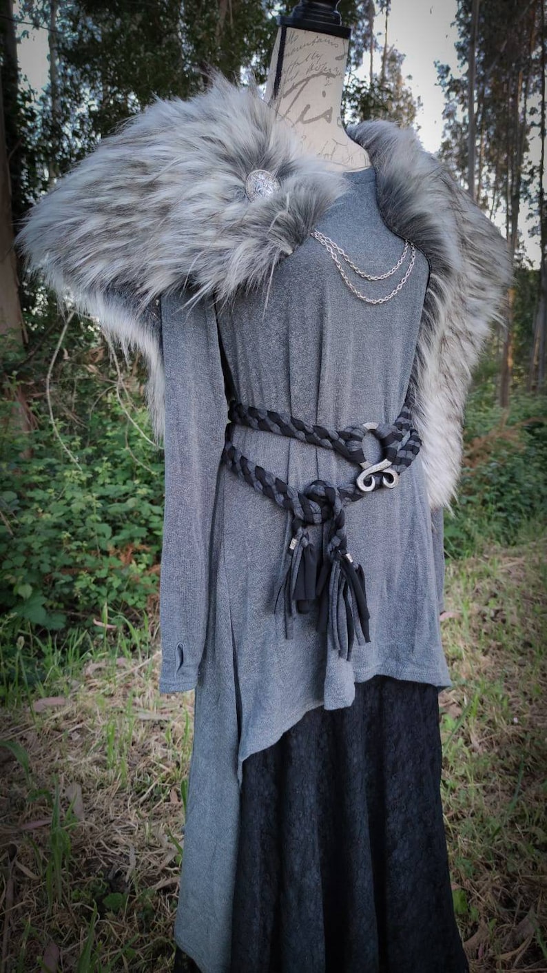 May include: A gray long-sleeved tunic with a black braided belt and a silver buckle. The tunic has a faux fur collar with a silver brooch. The model is wearing a black skirt underneath the tunic.