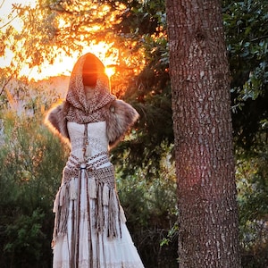Könnte beinhalten: Ein cremefarbenes böhmisches Hochzeitskleid mit Kapuzenschal und Fellschulterakzenten. Das Kleid hat komplizierte Spitzen- und Fransen Details, mit einem braunen gewebten Gürtel. Der Hintergrund zeigt einen Sonnenuntergang durch Bäume.