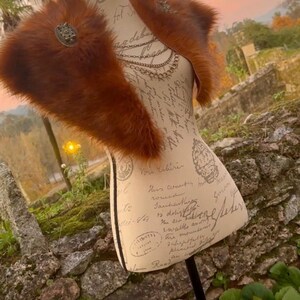 May include: A brown faux fur shawl with a decorative metal pin. The shawl is displayed on a mannequin with a vintage-style printed torso.