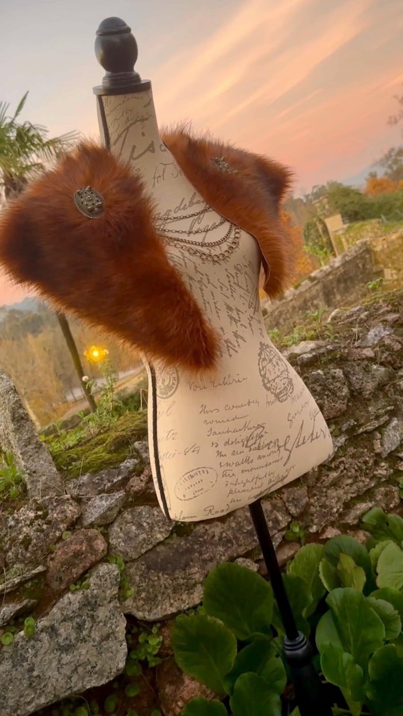 May include: A brown faux fur shawl with a decorative metal pin. The shawl is displayed on a mannequin with a vintage-style printed torso.