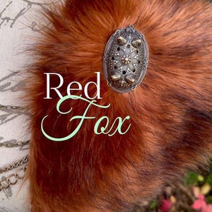 May include: A red fox fur collar with a silver and bronze ornate brooch. The collar is made of faux fur and has a soft, fluffy texture. The brooch is a decorative piece with intricate details.