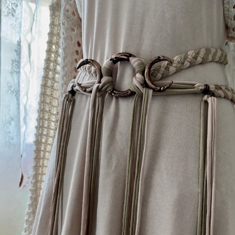 May include: A beige woven belt with a silver crescent moon buckle and long tassels. The belt is tied around a white dress.