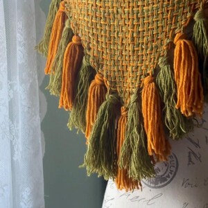 May include: A triangular woven shawl with a fringe of tassels in shades of green and orange. The shawl is displayed on a mannequin with a vintage-style fabric covering.