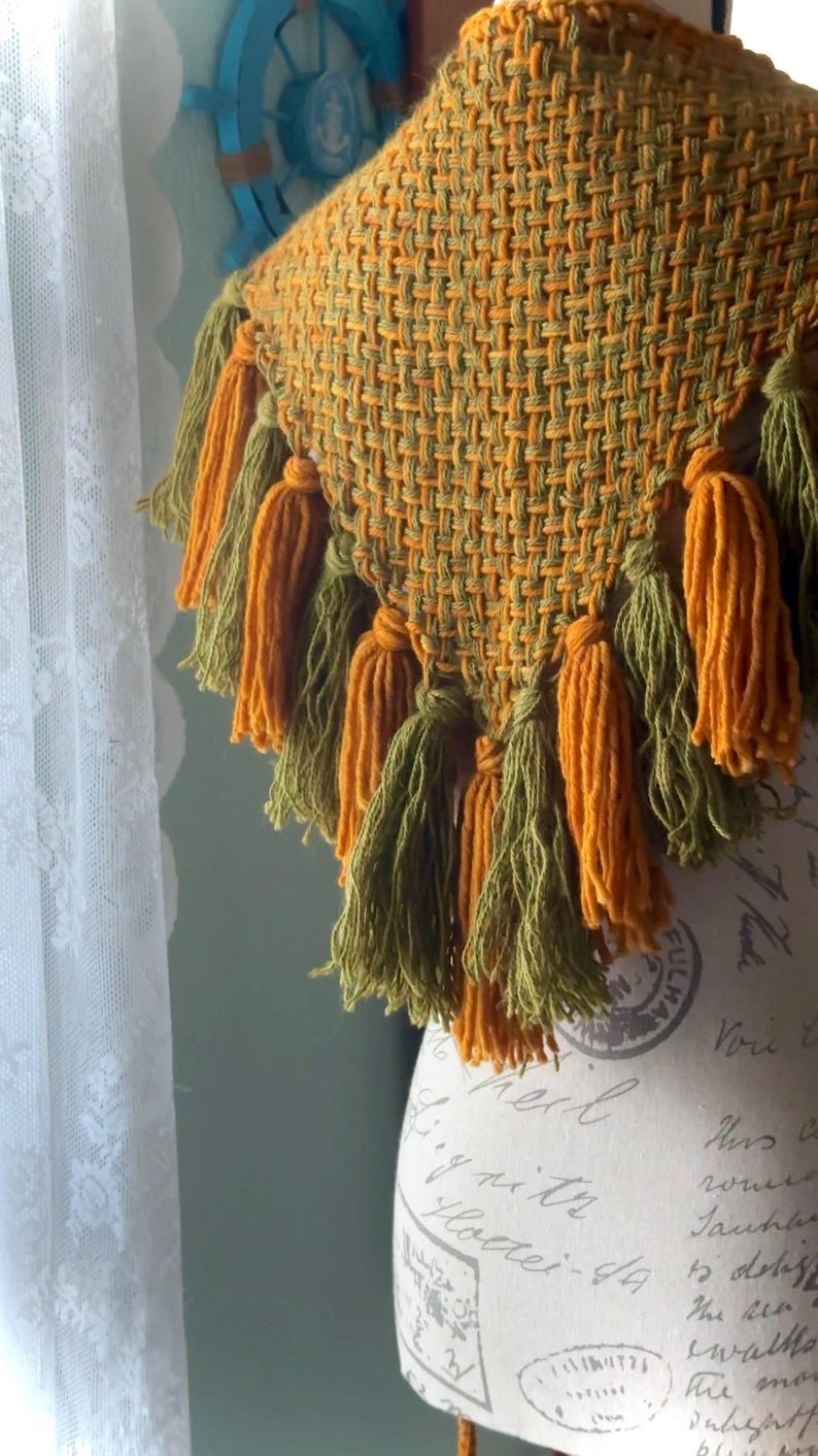 May include: A triangular woven shawl with a fringe of tassels in shades of green and orange. The shawl is displayed on a mannequin with a vintage-style fabric covering.