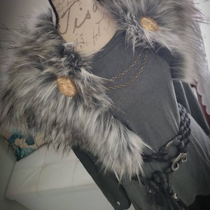May include: A gray fur collar with two gold buttons and a chain necklace. The collar is worn over a gray long-sleeved shirt with a black belt.
