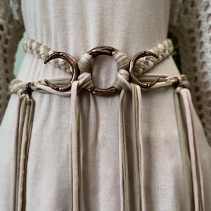 May include: A woven belt with a triple moon charm. The belt is made of three different colors of fabric: pink, green, and beige. The belt has long tassels that hang down from the bottom.