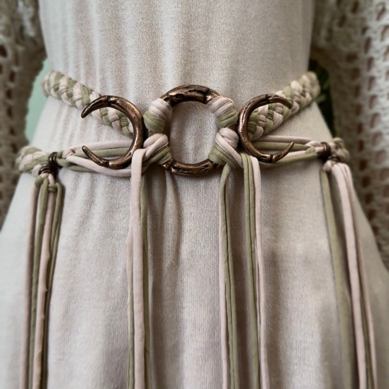 May include: A woven belt with a triple moon charm. The belt is made of three different colors of fabric: pink, green, and beige. The belt has long tassels that hang down from the bottom.
