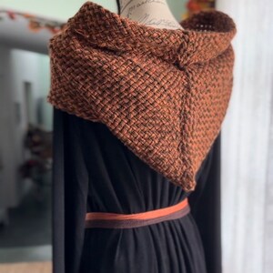 May include: A brown and black knitted hooded scarf with a woven texture. The scarf is draped over a black dress with a brown belt.