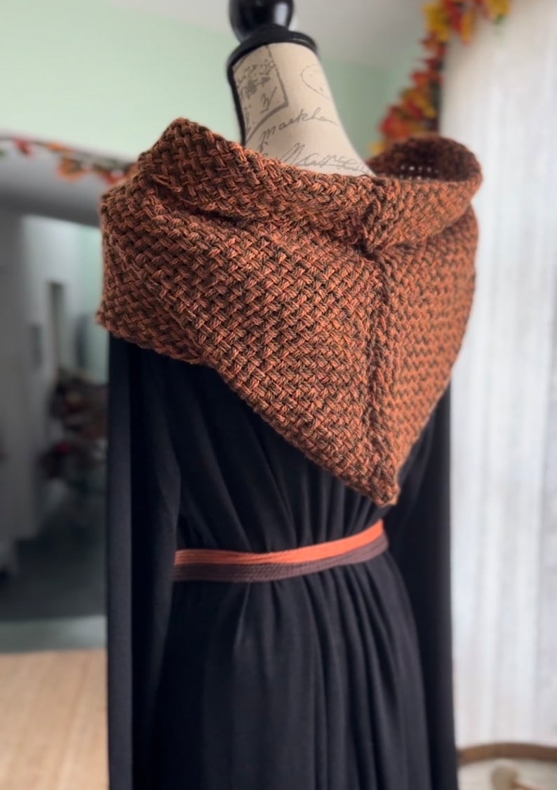 May include: A brown and black knitted hooded scarf with a woven texture. The scarf is draped over a black dress with a brown belt.