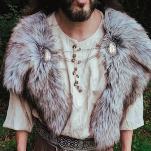 May include: A person wearing a white shirt with a fur collar and silver accents. The collar is fastened with two decorative silver buckles. The shirt has a chain necklace with silver accents.