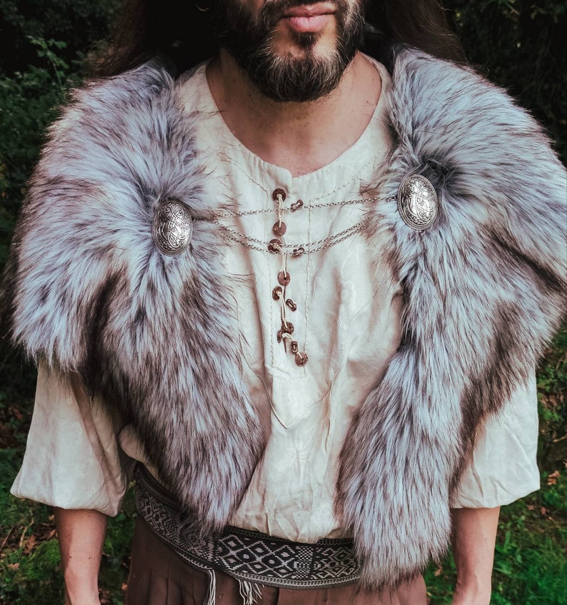 May include: A person wearing a white shirt with a fur collar and silver accents. The collar is fastened with two decorative silver buckles. The shirt has a chain necklace with silver accents.
