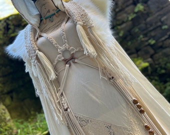 May include: Cream-colored hooded cloak with faux fur trim and macrame detailing. The cloak features a long, flowing design with intricate rope and wooden bead accents. A cream-colored dress is visible underneath, with a lace panel and a fabric flower.